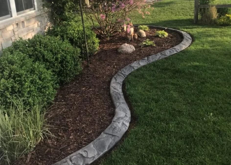 Curved garden bed with stone edging, lush green bushes, and a pink-blooming shrub, set against a well-maintained lawn, creating a serene landscape.
