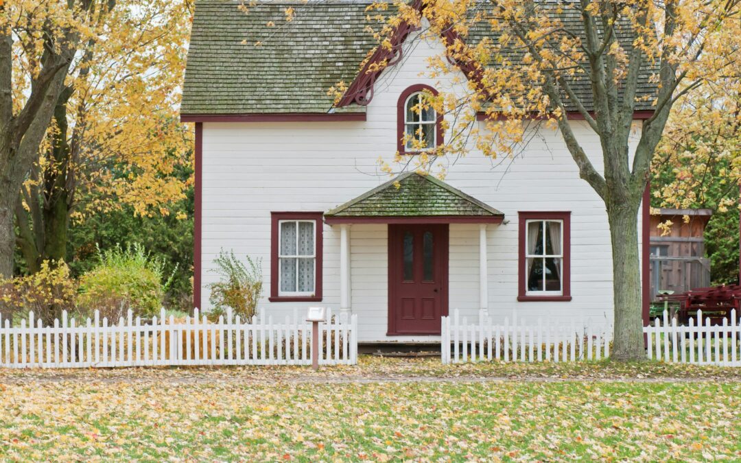 Charming white house with red accents, set amidst autumn foliage. A white picket fence surrounds the yard, covered in fallen leaves, evoking a cozy, nostalgic feel.