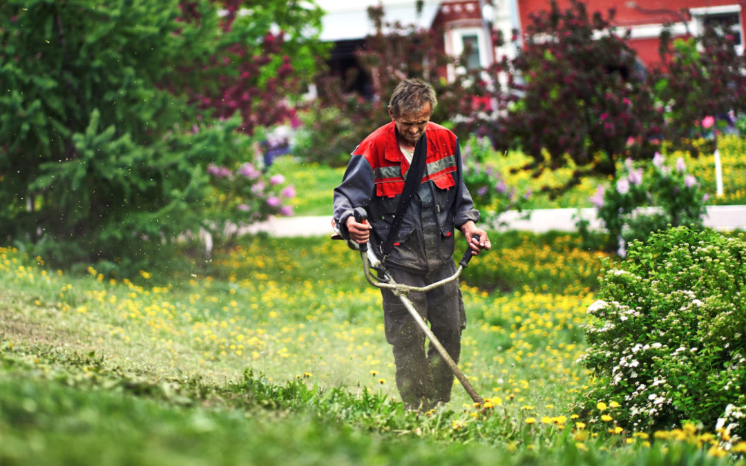A person in a red and gray jacket mows grass with a trimmer in a garden filled with green trees and colorful blooming flowers, conveying a sense of tranquility.