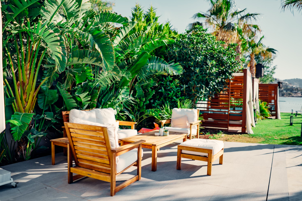 Cozy patio scene with wooden chairs and a table on a sunlit terrace, surrounded by lush green plants. The atmosphere is tranquil and inviting.
